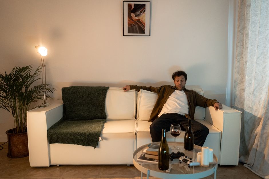 An adult man looking stressed, seated on a sofa with wine bottles on the table.