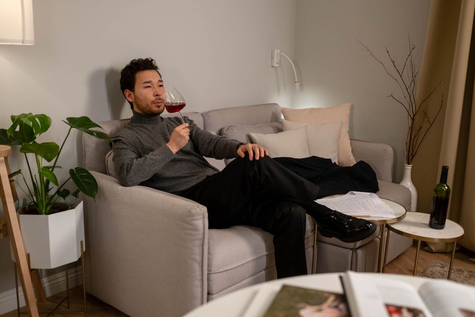 Asian man relaxing at home, enjoying a glass of red wine while sitting on a comfortable sofa.