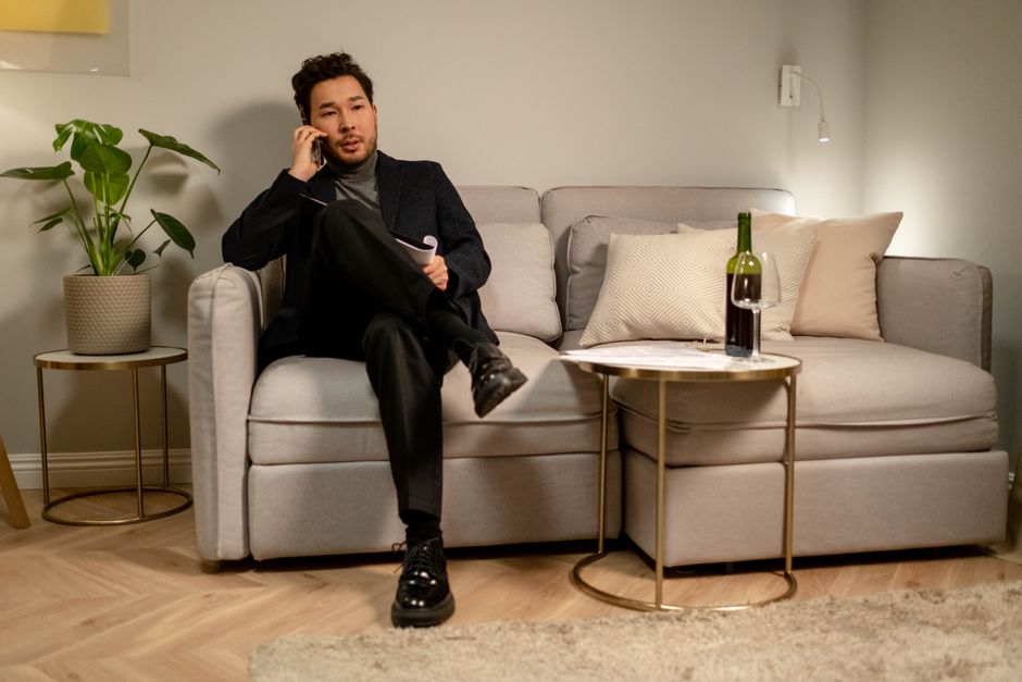 Asian man sitting on a sofa, talking on phone, with a wine bottle and glass on a side table.