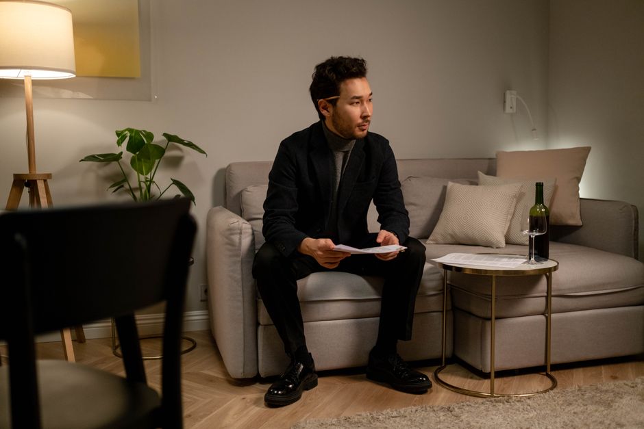 A man sits on a sofa reading papers with a wine bottle on a table in a warmly lit room.